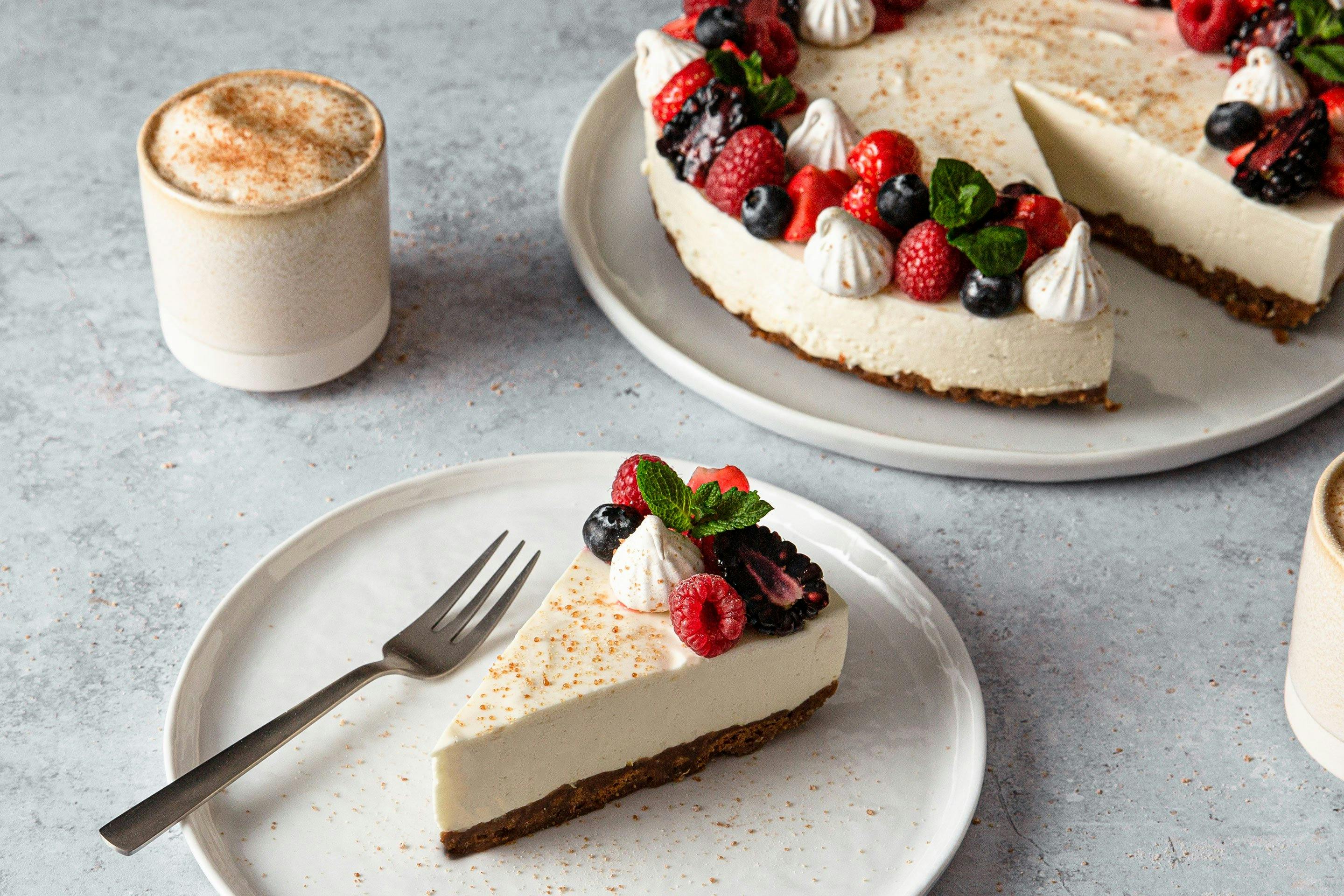 Cheesecake mit frischen Beeren garniert auf einer Kuchenplatte. Im Vordergrund ein Stück Cheesecake auf einem Teller mit Kuchengabell und einem Cappuccino in einem Becher.