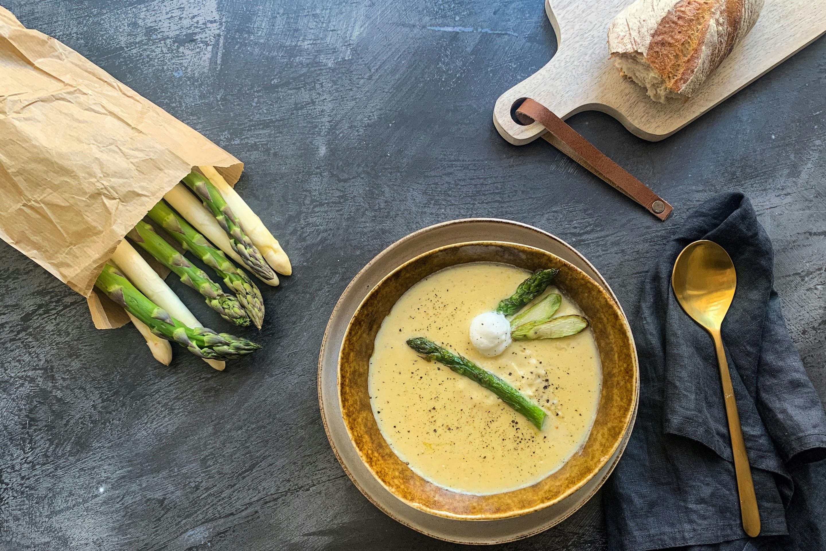 Spargelcreme-Suppe mit grünem Spargel in einem tiefen Teller; daneben liegt ein Stück Baguette und Spargel