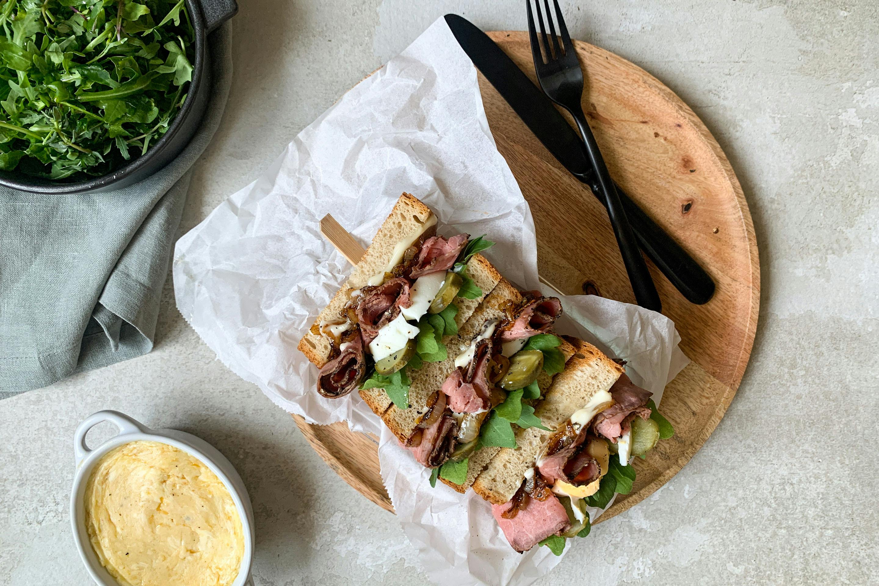 Roastbeef-Sandwich auf Butterbrotpapier auf einem Holzbrett mit schwarzem Besteck und Sauce in einem Schälchen und einem grünen Beilagensalat