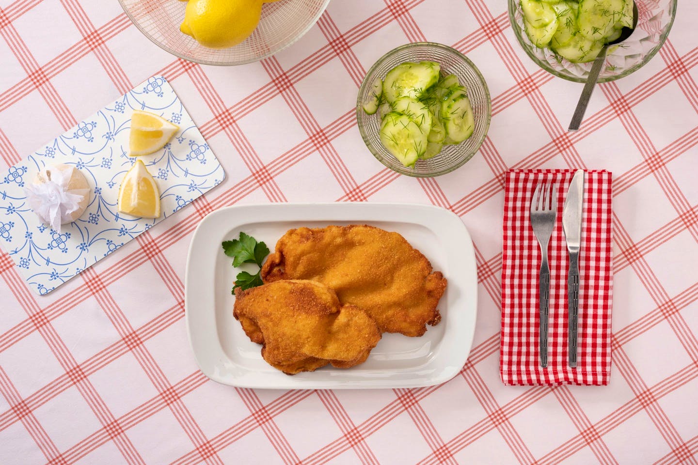 2 Wiener Schnitzel auf einem rechteckigen Teller, mit Gurkensalat in einem gläsernen Schälchen, auf rot-weiß-karierter Tischdecke