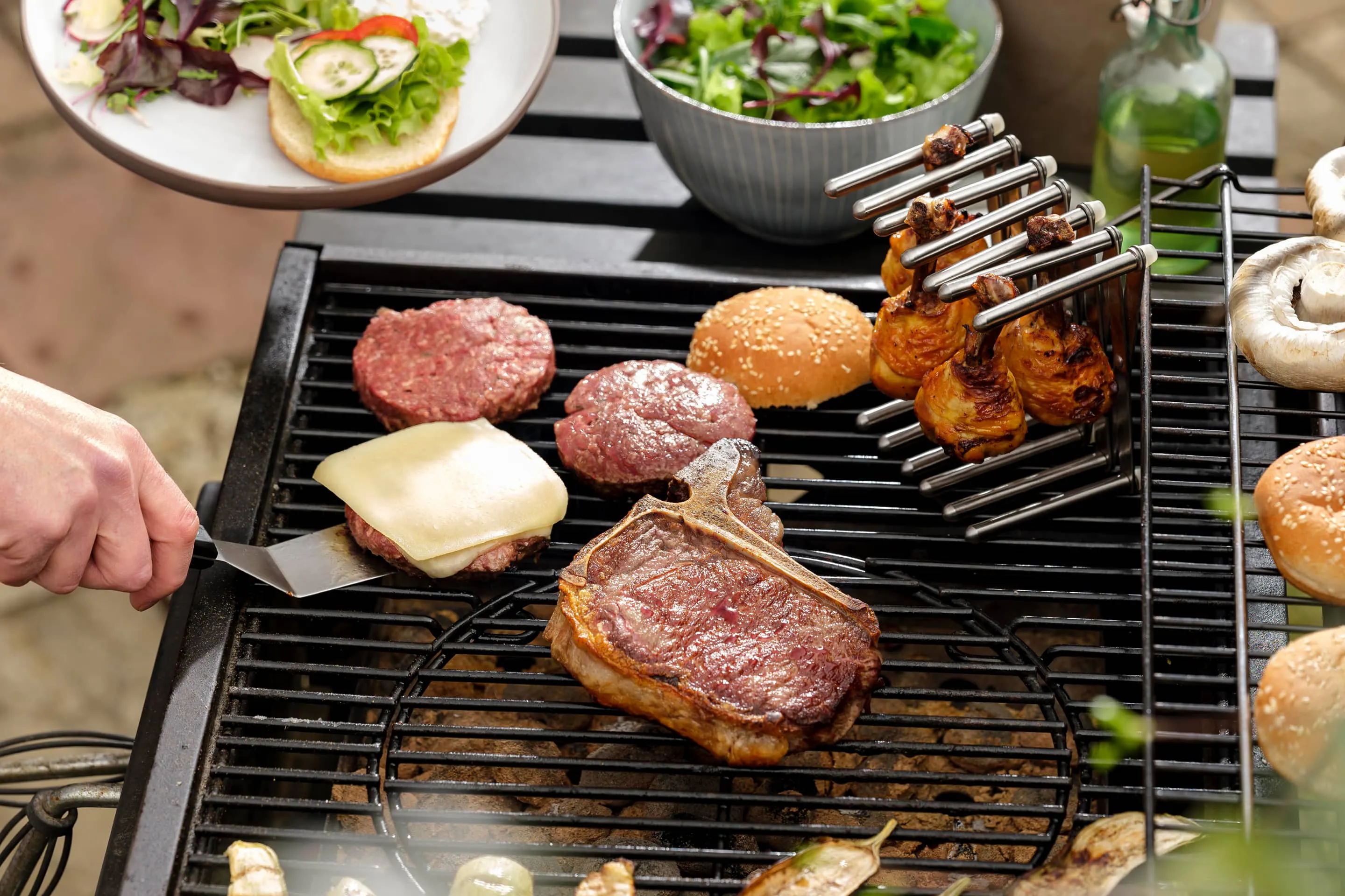 Burger Patties, Chicken Drumsticks und Steak auf dem Grill.